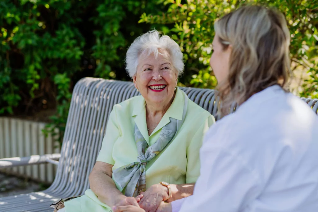 Elderly woman with white hair wearing light green blouse laughing while holding hands with caregiver on outdoor bench, surrounded by greenery