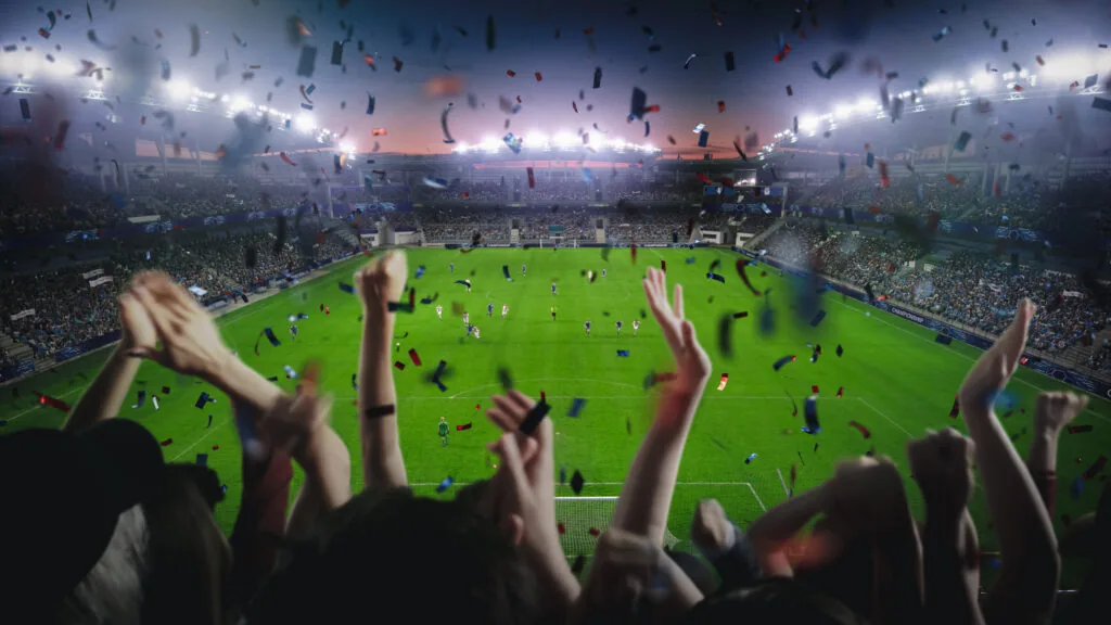 Fans cheer and throw confetti in a packed, brightly lit stadium during a nighttime soccer match, with players visible on the field and the stands full of spectators.